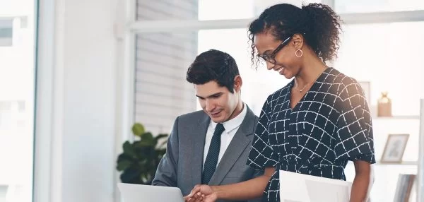 Two professionals, one in a suit and one in a patterned dress, stand together in an office, smiling at a laptop as they collaborate on Sage Intacct Best Practices for Nonprofits.
