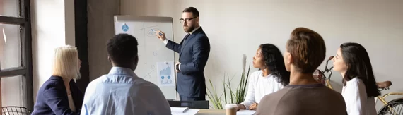 A man in a suit presents charts on a flip chart, discussing nonprofit streamlining process and maintaining internal controls to a group of four colleagues seated around a table in a modern office meeting.