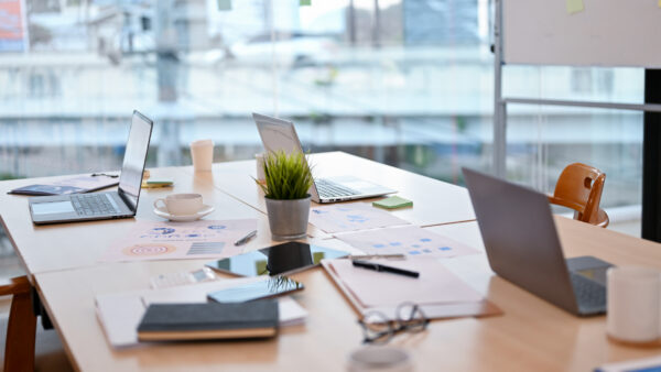 A modern office meeting table set for a quarterly estimated tax payment discussion features open laptops, documents, notebooks, coffee cups, and a potted plant. Bright natural light streams in from large windows in the background. No people are present.