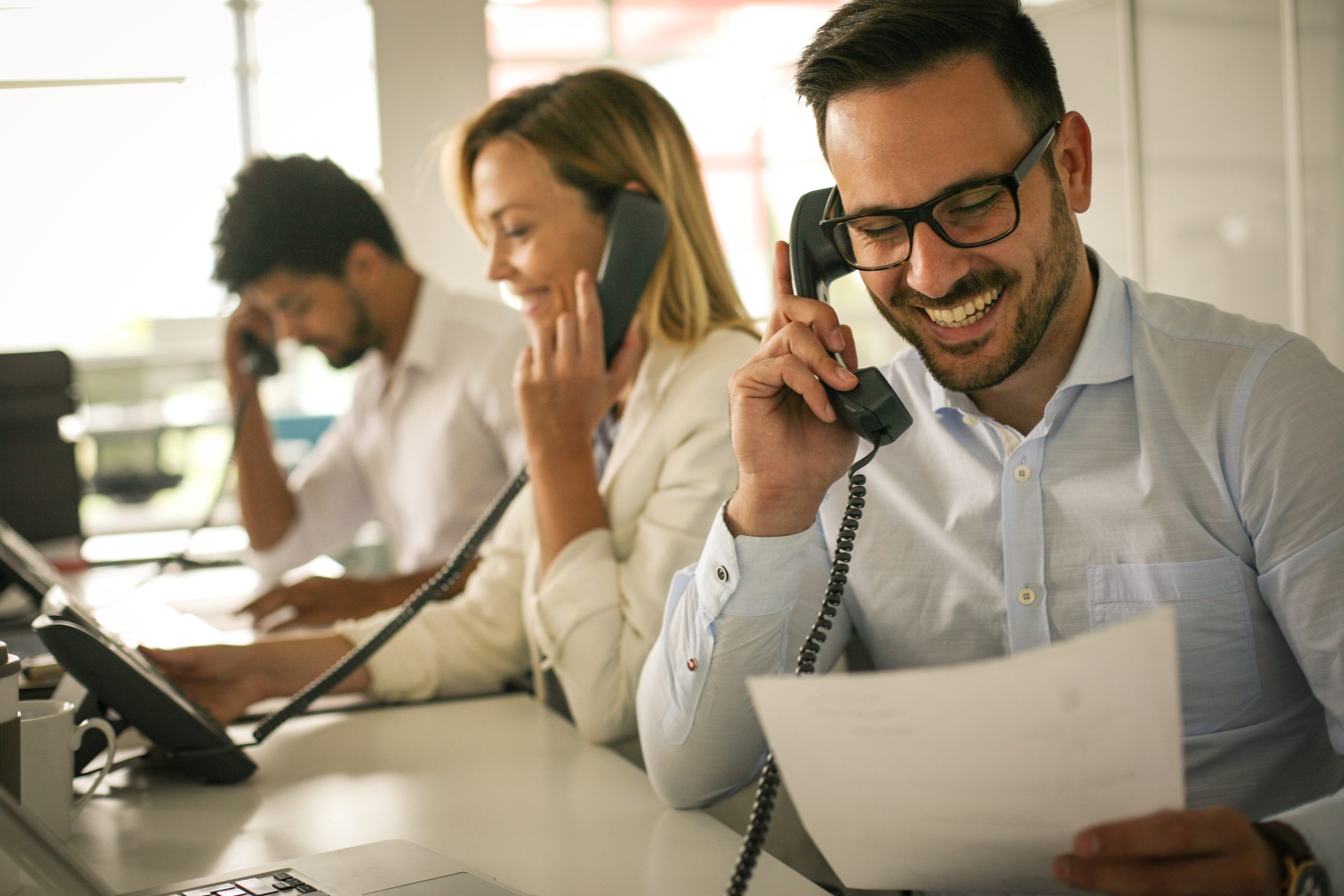 Three office workers sitting at desks, smiling and talking on corded phones. The man in the foreground is holding a document related to payroll tax compliance, while the others are focused on their calls and computers. The setting is bright and professional.