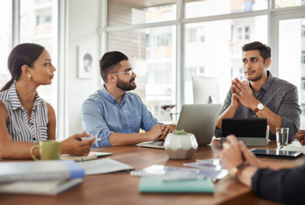Four people sit around a table in a modern office, engaged in a serious discussion. Laptops, notebooks, and a plant are on the table. Large windows in the background let in natural light.