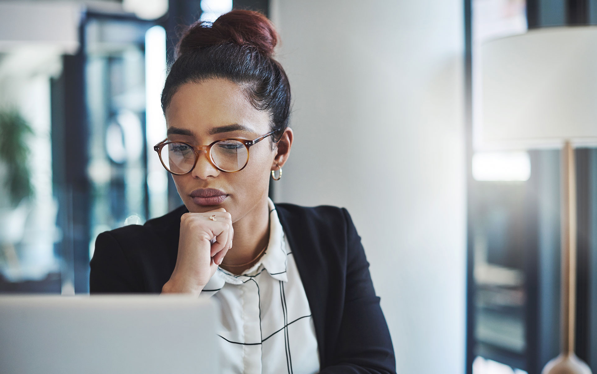 A woman wearing glasses and business attire sits at a desk, looking thoughtfully at her laptop screen with her hand resting on her chin. She appears focused and is in a modern office setting.