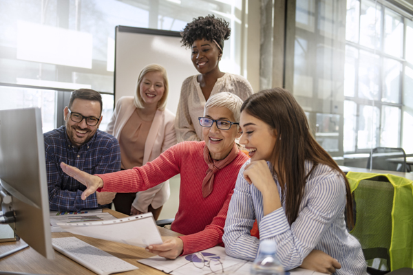A group of five diverse colleagues smiling and collaborating at a desk in a bright, modern office. One woman points at a computer screen displaying ira elective pay documents, while others look on and engage with interest.