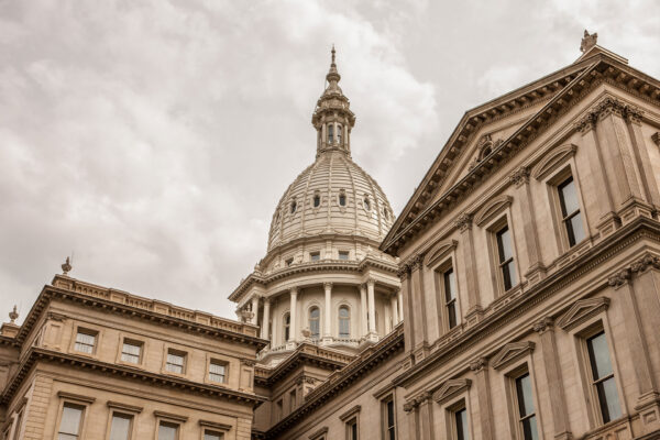 The image shows the ornate dome and upper facade of a historic government building, featuring classical architectural details and beige stonework against a cloudy sky.