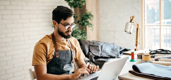 A man wearing glasses and a leather apron sits at a desk working on a laptop. There are sewing materials and fabrics on the table, and a lamp and plant in the background.