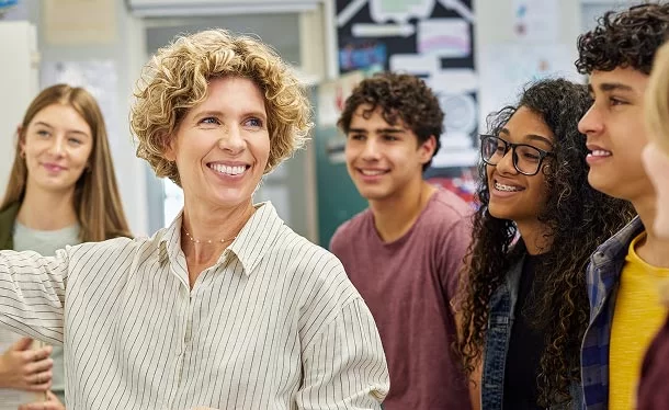 A teacher with curly hair smiles and stands in front of a group of diverse high school students in a classroom, engaging them in conversation or a lesson. The students look happy and attentive.