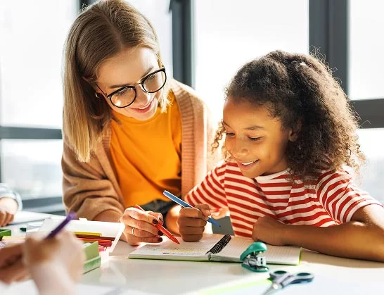 A young girl in a red striped shirt smiles while doing schoolwork, as an adult woman with glasses and a warm sweater sits beside her, helping and pointing at her notebook in a bright classroom.
