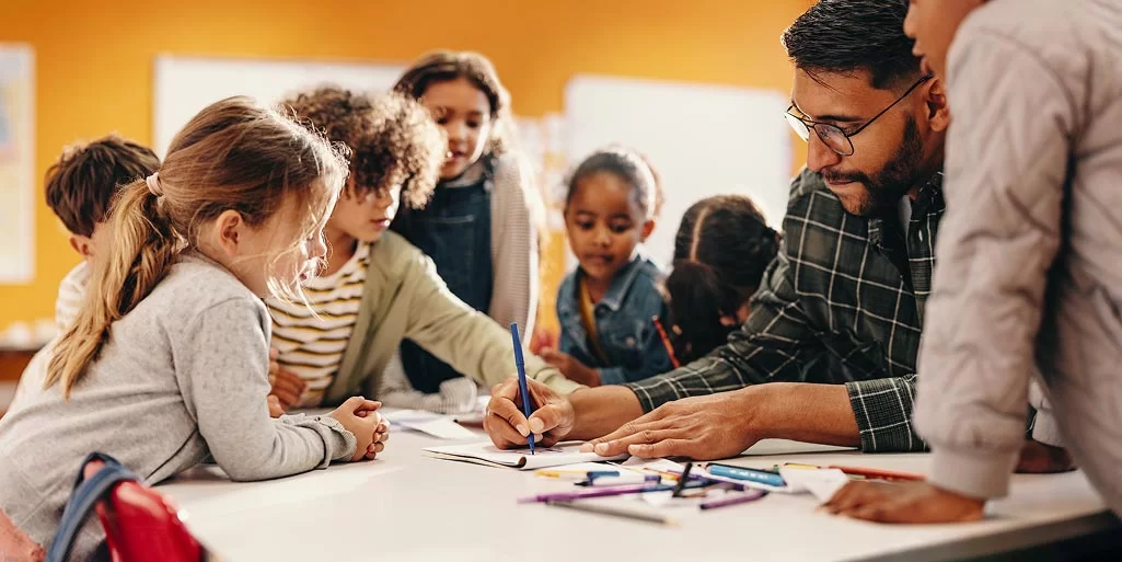 A teacher helps a group of young children with a drawing at a table, surrounded by colored pencils, in a bright classroom setting.
