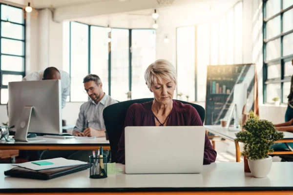 A woman with short blonde hair works on a laptop at a desk in a modern, bright office. Two men are working in the background, and potted plants and office supplies are on the desk. Sunlight streams through large windows.