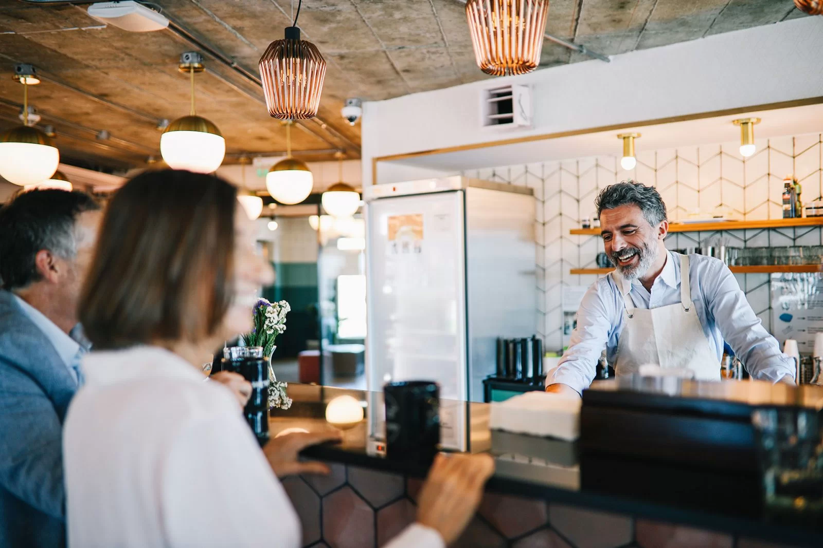 A smiling barista with gray hair and a white apron chats with two customers at a modern coffee shop counter, surrounded by hanging lights and stylish decor.