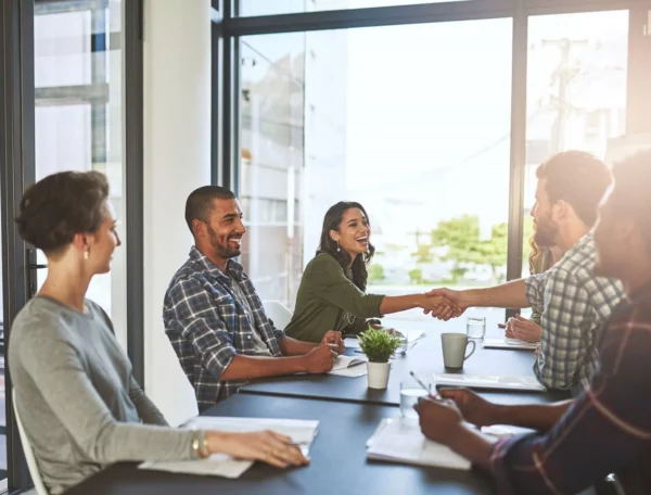 A group of people sit around a conference table in a bright office. Two people, a woman and a man, smile and shake hands across the table, while others watch and smile. Papers and a small plant are on the table.