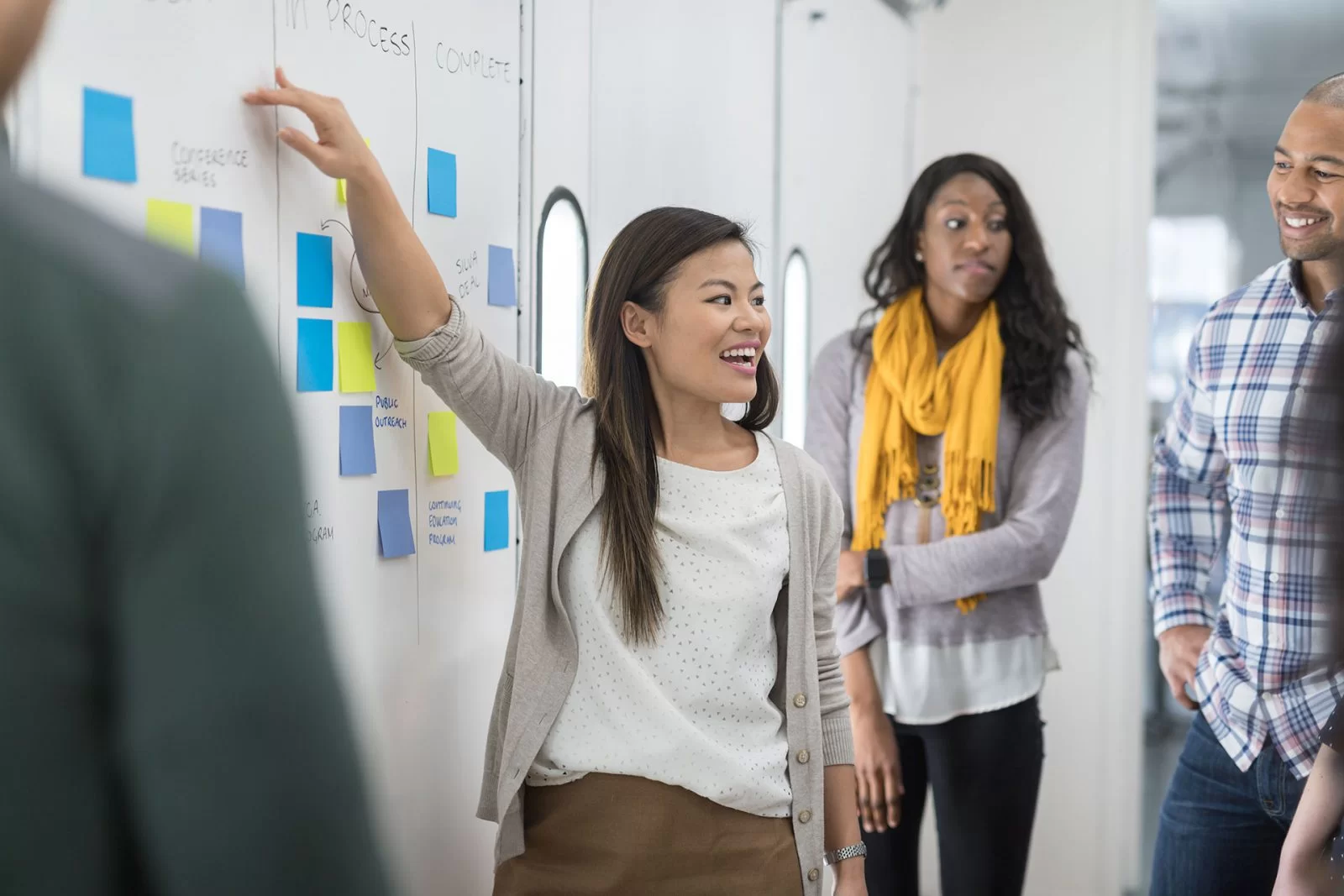 A woman stands by a whiteboard covered with sticky notes, smiling and gesturing while talking to three colleagues. The group appears engaged in a discussion in a modern office setting.