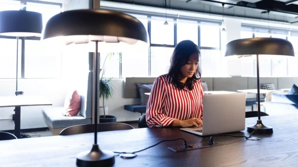 A woman in a red and white striped shirt sits at a desk in a modern, well-lit office space, working on a laptop beneath large desk lamps.