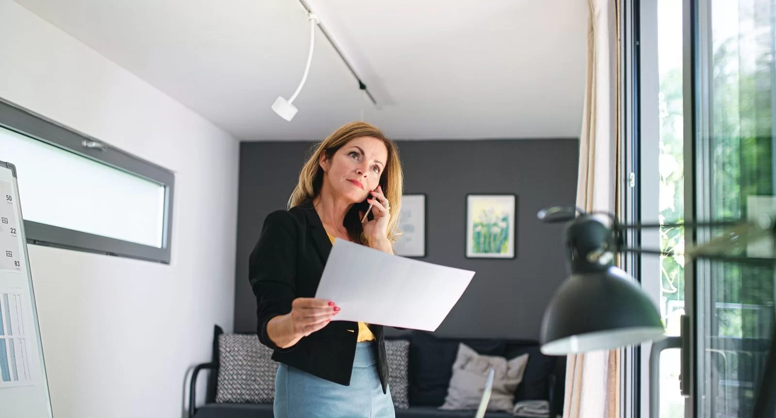 A woman stands in a modern office, talking on her phone while holding a sheet of paper. She looks thoughtful, with natural light coming in through large windows beside her.