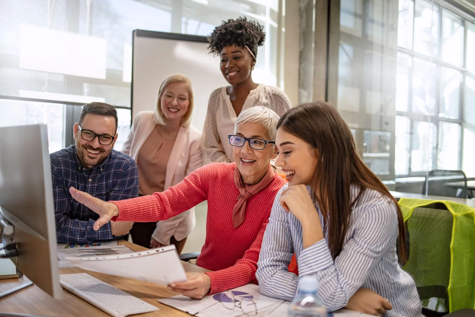 Five colleagues smile and look at a computer screen in a bright office. A woman in a red sweater points at a paper while others gather around, appearing engaged and collaborative.