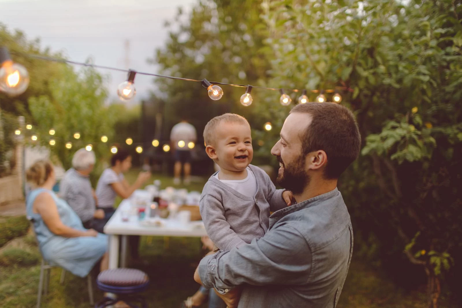 A man smiles as he holds a laughing baby outdoors during a garden gathering. String lights hang above, and several people sit at a table with food in the background, surrounded by greenery.