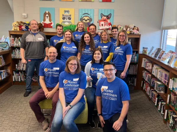 A group of thirteen people wearing matching blue Maner Costerisan Cares t-shirts pose together in a library, surrounded by bookshelves and stuffed animals with animal art on the wall behind them.