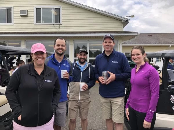 Five adults pose and smile outdoors in front of a light-colored building, dressed in casual golf attire with jackets and caps. Some hold drinks, and golf carts are visible in the background.