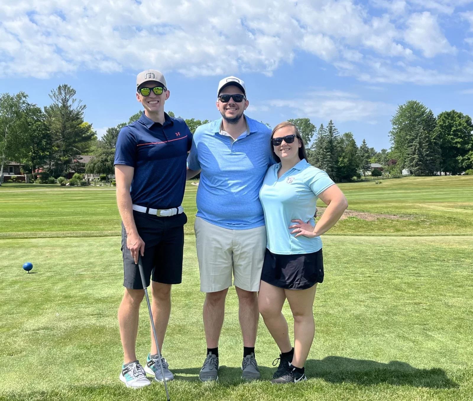 Three people stand smiling on a golf course under a partly cloudy sky. Two men and one woman pose together; one man holds a golf club. All are wearing sunglasses and casual golf attire. Trees and houses are visible in the background.