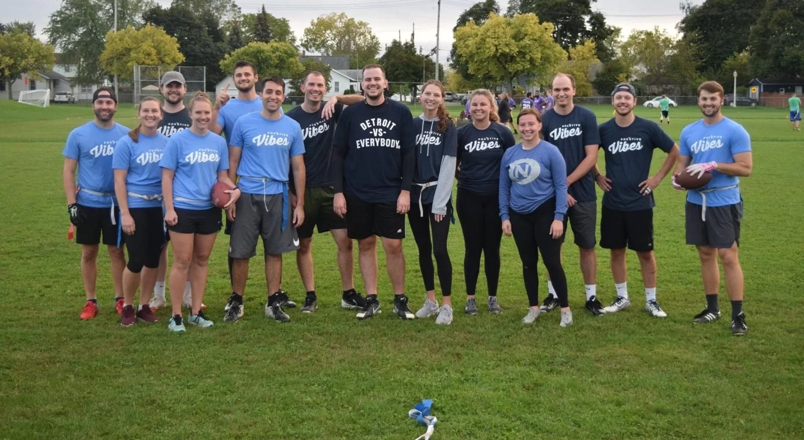 A group of 15 people, some holding footballs, stand smiling on a green field. Most are wearing athletic clothing and matching Vibes shirts. Trees and more people are visible in the background.