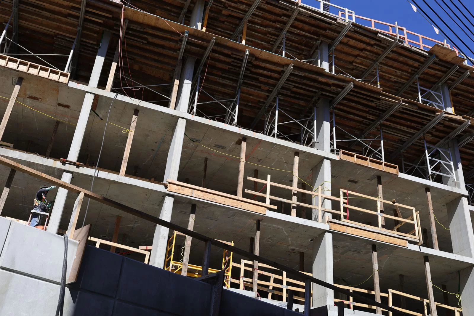 A multi-story building under construction with exposed concrete beams, scaffolding, and wooden planks. A construction worker in safety gear is visible on the lower left side. Blue sky is in the background.