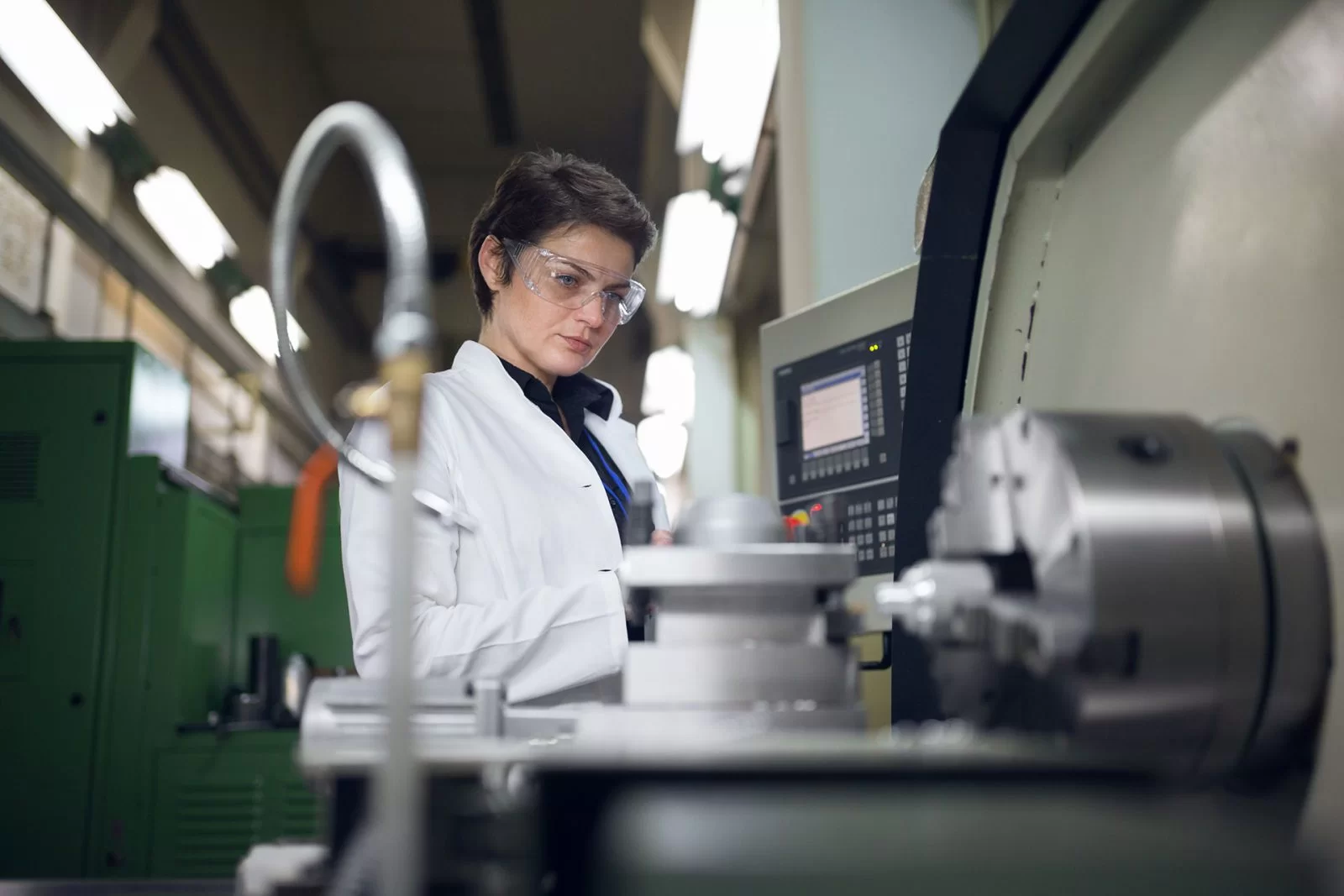 A person wearing safety glasses and a lab coat operates a CNC machine in an industrial or laboratory setting, focusing on the control panel, with machinery in the foreground.