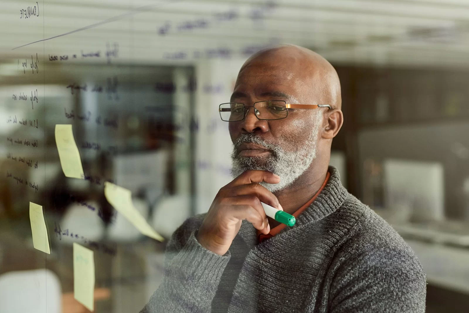 A man with glasses and a beard thoughtfully looks at notes and writing on a glass wall, holding a green marker and resting his hand on his chin in a modern office setting.
