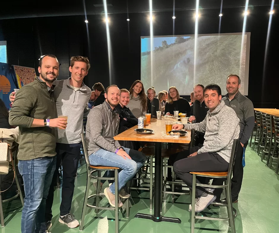 A group of people sit and stand around a high table in a large indoor space, smiling at the camera. Drinks and food are on the table, and a large screen displaying a nature scene is visible in the background.