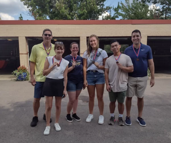 Six people stand outside wearing medals around their necks. Some are holding up one finger in a “number one” gesture. They are smiling and appear to be celebrating an achievement or event on a sunny day.
