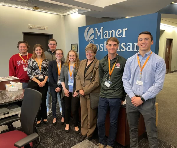 Eight people stand and smile in an office lobby in front of a blue wall with the Maner Costerisan logo. Most wear business attire and name badges with orange lanyards. Desks and office furniture are visible.