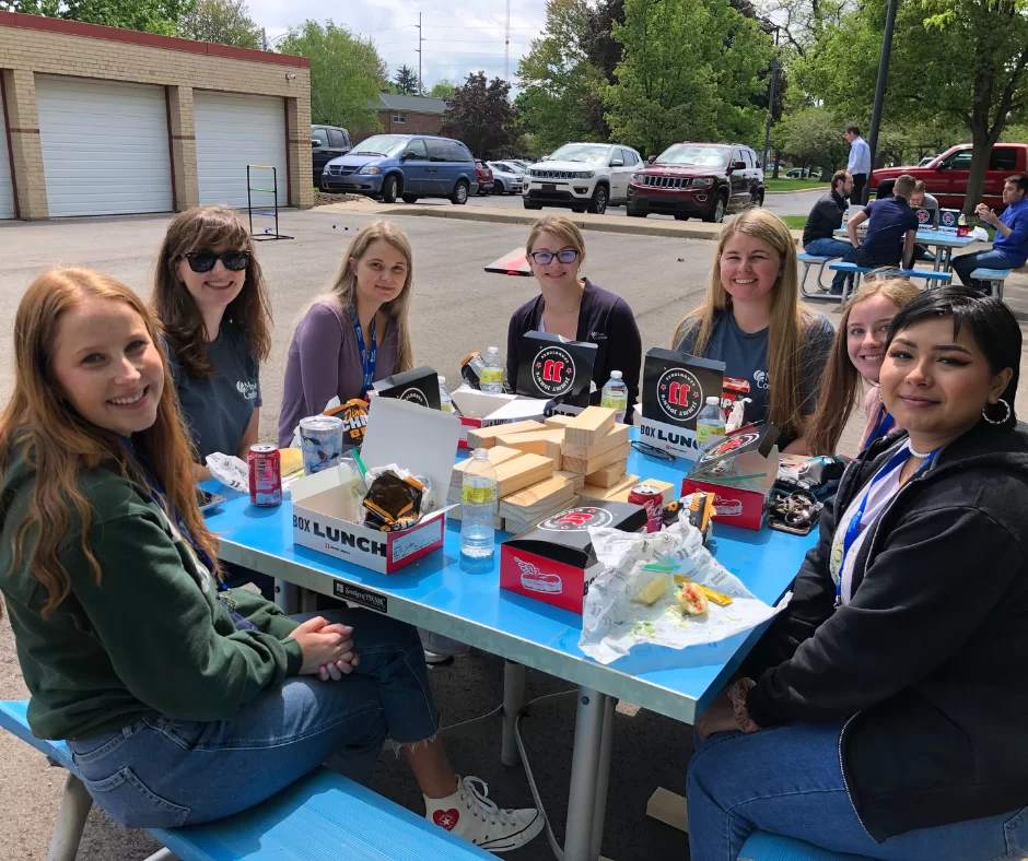 Seven young women sit around a blue picnic table outside, smiling and enjoying boxed lunches on a sunny day. The background shows trees, parked cars, and other people at picnic tables.