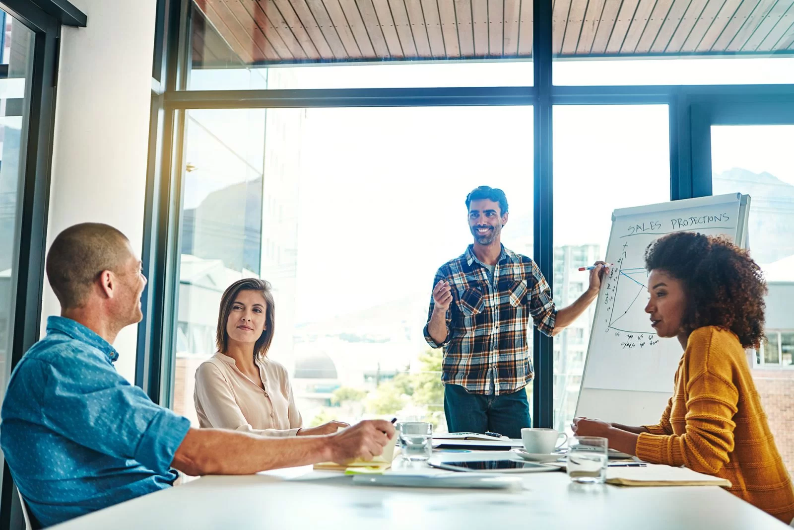 A man stands by a flip chart giving a presentation to three colleagues seated at a table in a bright office with large windows. Two women and one man listen and take notes.