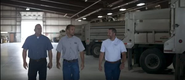 Three men in work shirts and jeans walk together inside a large industrial garage, with two large trucks parked nearby and natural light coming through the open door.