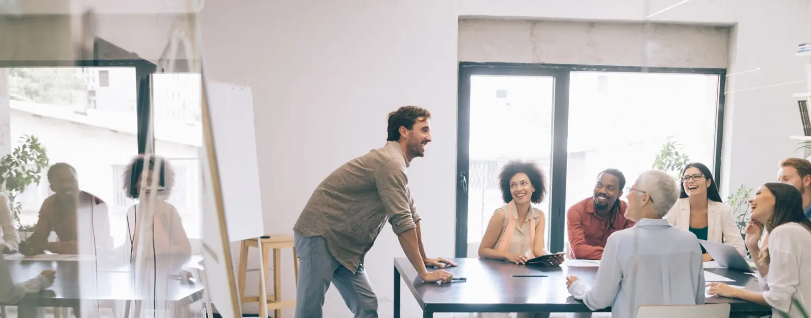 A man stands smiling and talking to a group of six people seated around a table in a bright office, all appearing engaged and happy during a meeting. Large windows let in natural light.