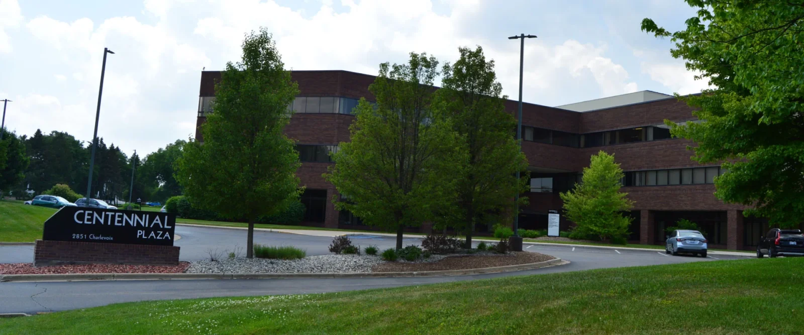 A brick office building labeled Centennial Plaza is surrounded by trees and landscaping, with parked cars and a driveway leading to the entrance on a partly cloudy day.