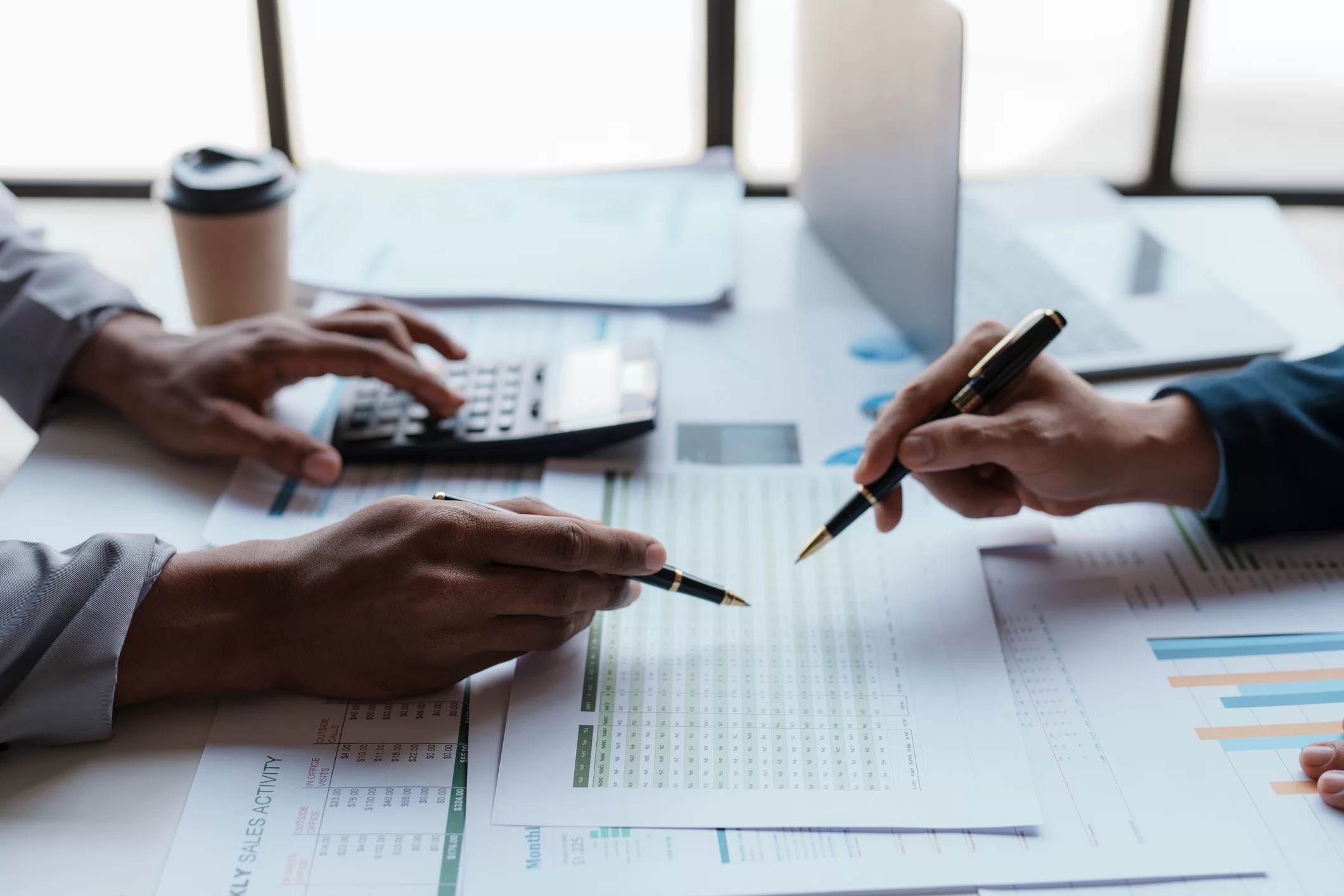 Two people sit at a desk reviewing documents and graphs, with one using a calculator and both holding pens, suggesting a business meeting or financial discussion. A laptop and a coffee cup are also on the table.