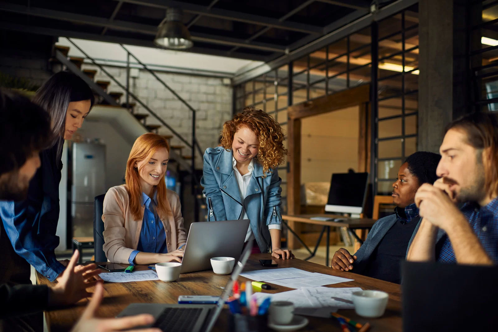 A diverse group of six people sit and stand around a table in a modern office, smiling and collaborating over a laptop and documents during a meeting.