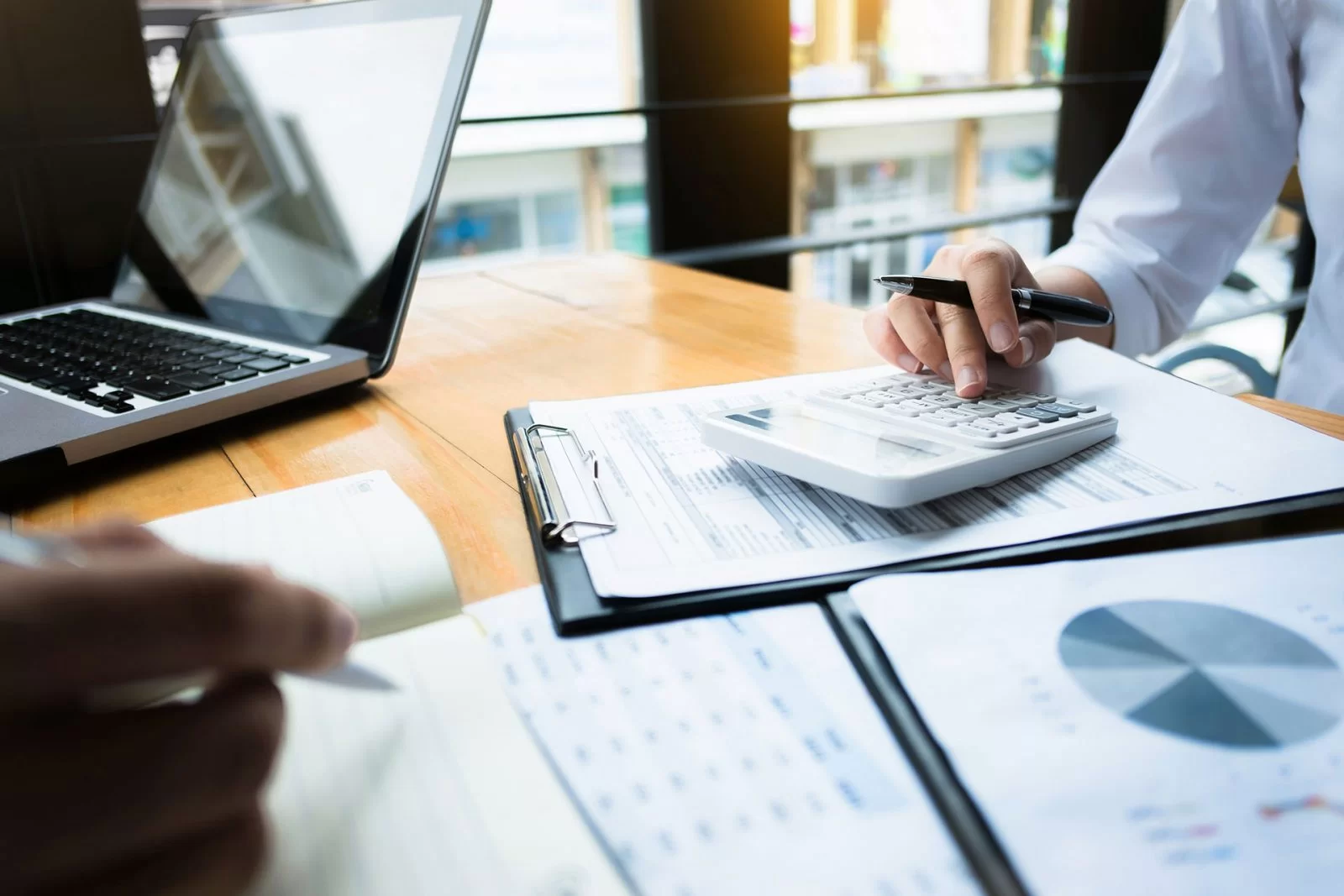 Two people work at a desk with documents, a laptop, and a calculator. One person types on the calculator, while the other writes in a notebook. A financial report with charts is visible on the table.
