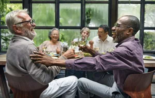 Two older men sit at a table, laughing and clinking glasses of white wine, while three other people in the background smile and enjoy a meal together in a bright, modern setting.