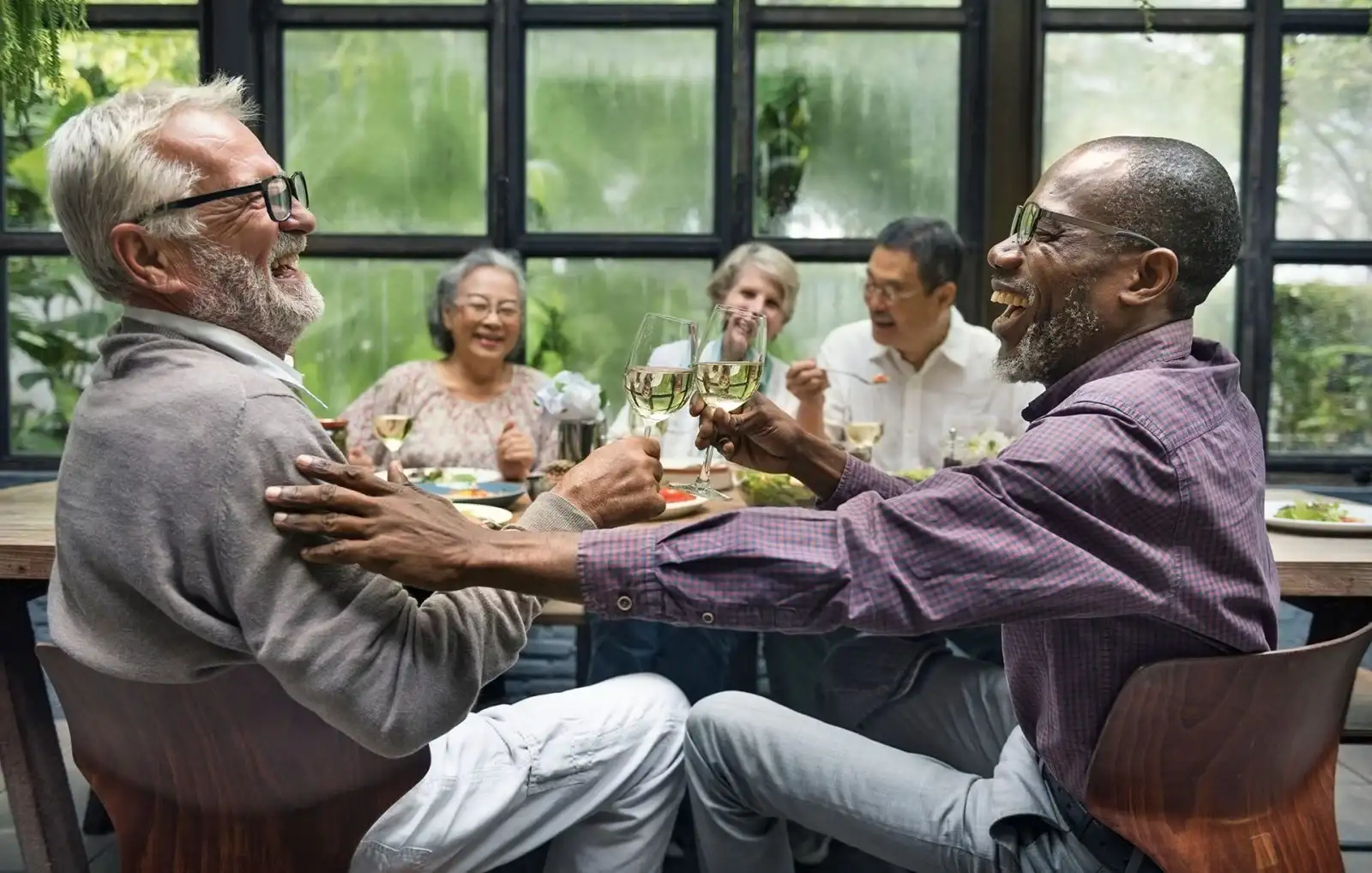 Two older men sit at a table, laughing and clinking glasses of white wine, while three other people in the background smile and enjoy a meal together in a bright, modern setting.