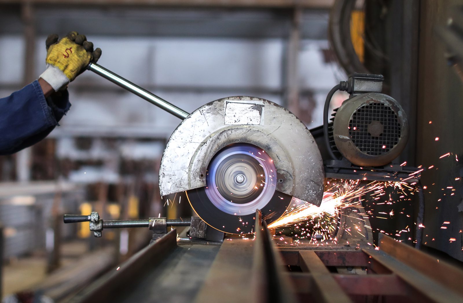 A close-up of a circular saw cutting through metal, producing bright sparks. A gloved hand holds the material steady, and machinery is visible in the background of an industrial workshop.