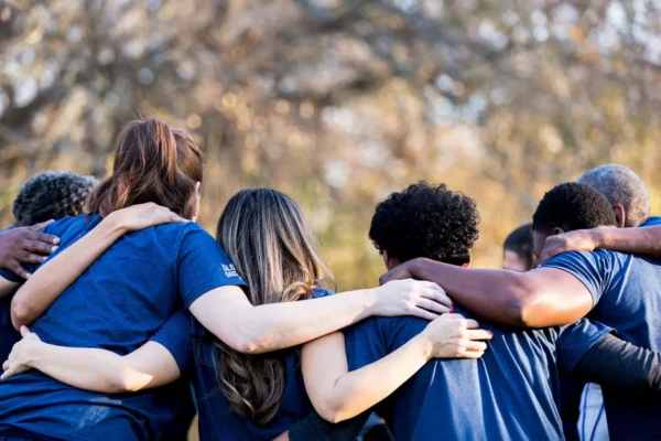 A group of people wearing blue shirts stand outdoors with their arms around each other’s shoulders, seen from behind, showing unity and teamwork. Trees with autumn leaves are blurred in the background.