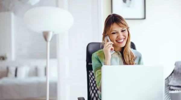 A smiling woman sits at a desk in a bright room, talking on her smartphone while looking at her laptop.