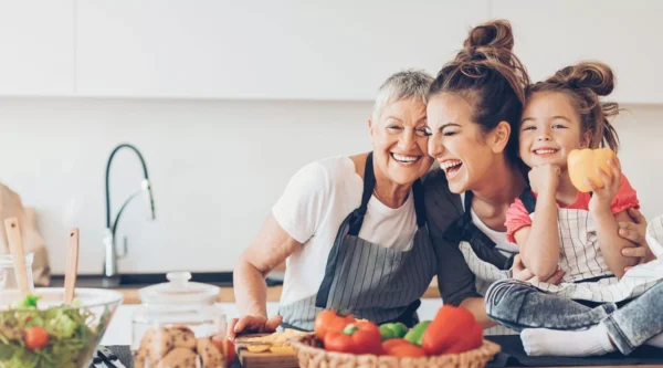 Three generations of women—an elderly woman, a young woman, and a child—smile and laugh together in a kitchen, surrounded by colorful vegetables on the counter. The child holds a yellow bell pepper.
