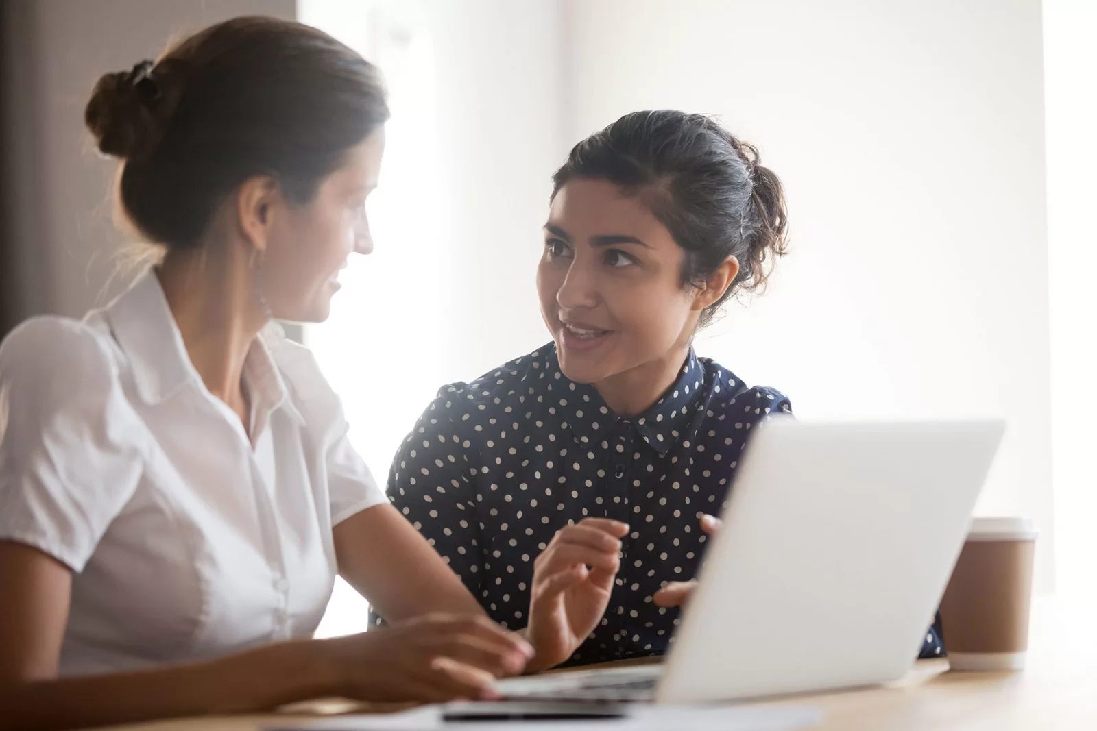 Two women sit at a table, engaged in conversation, with a laptop open in front of them. One woman wears a white shirt, the other a dark polka-dot blouse. A coffee cup is also visible beside the laptop.