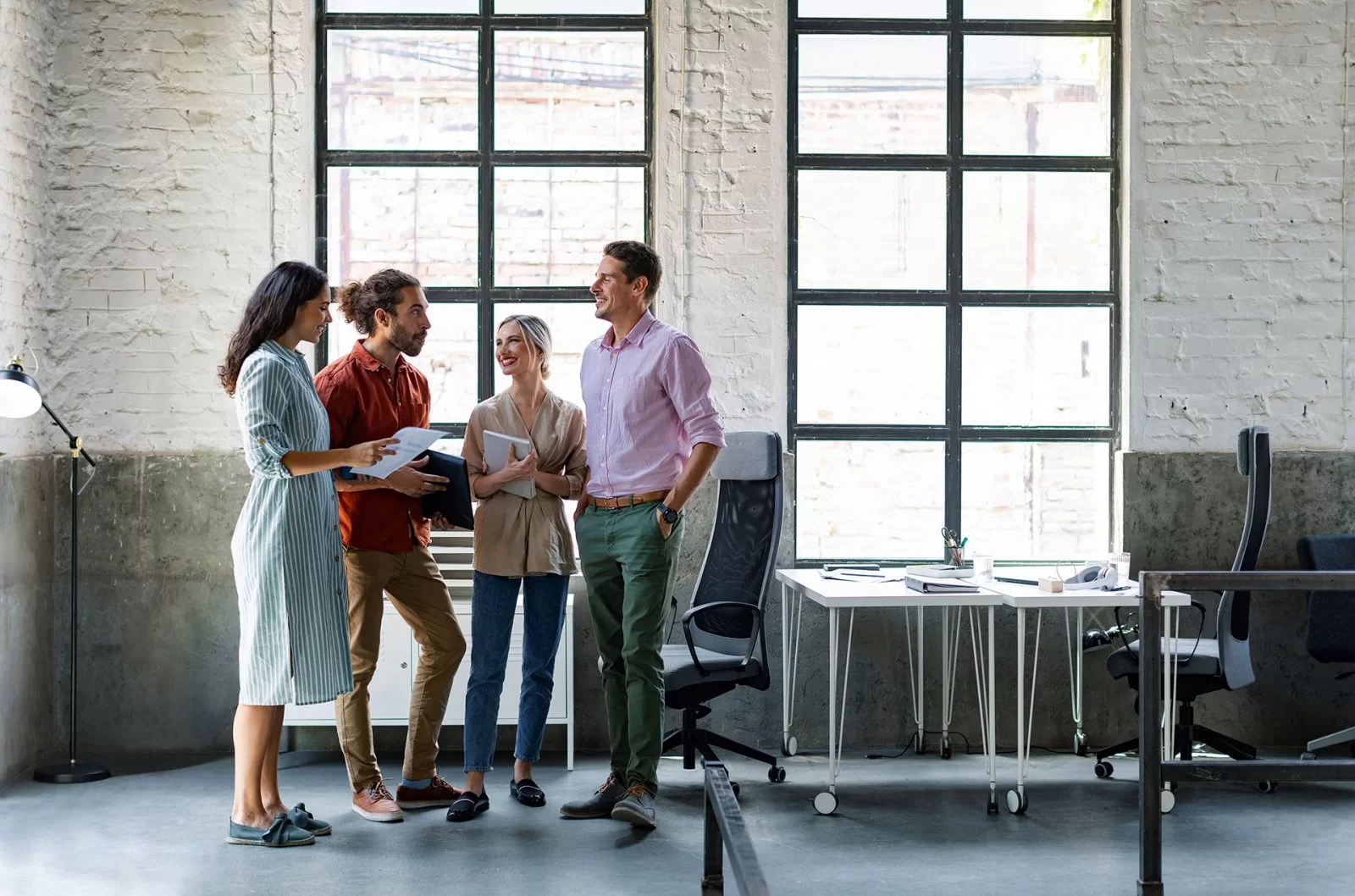Four people stand and talk in a modern, bright office with large windows. Two hold notebooks, one holds a laptop, and they appear to be having a casual discussion near a desk with office supplies.