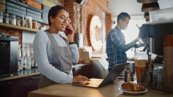 A smiling barista in glasses and an apron talks on the phone and uses a laptop at a coffee shop counter, while a coworker prepares a drink at an espresso machine in the background.