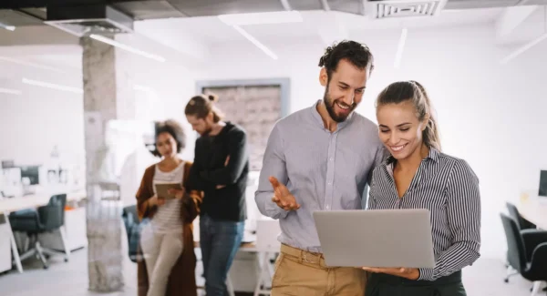 Two coworkers stand and smile while looking at a laptop in a modern office, with two other colleagues talking in the background. The office is bright, open, and filled with desks and computers.