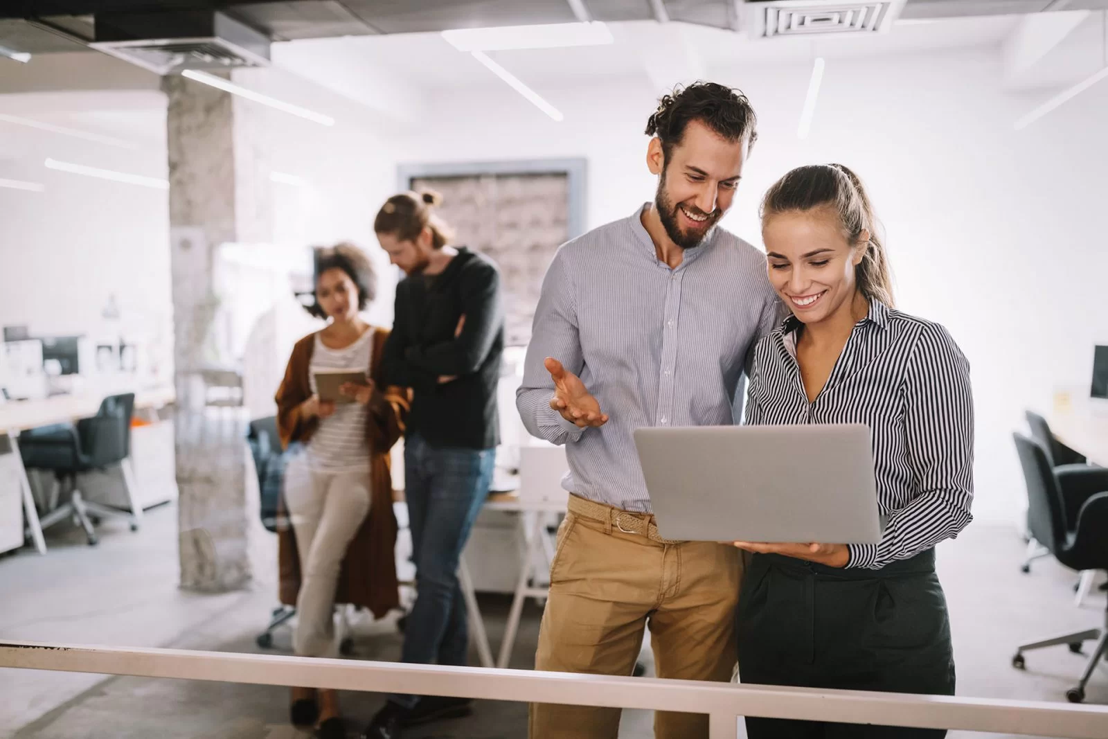 Two coworkers stand and smile while looking at a laptop in a modern office, with two other colleagues talking in the background. The office is bright, open, and filled with desks and computers.