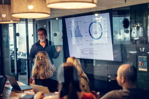A man stands in front of a screen displaying charts and graphs, presenting to a group of people seated around a conference table in a modern office meeting room.